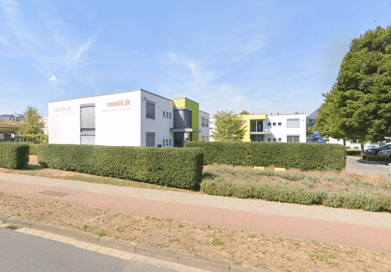 The image shows a modern office building with "reinstil.de" signage, surrounded by trimmed hedges and greenery, on a sunny day with a clear sky.