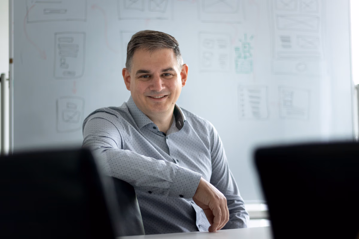 A man in a light gray shirt smiling in an office setting, with a whiteboard featuring sketches and diagrams related to inspection software in the background.